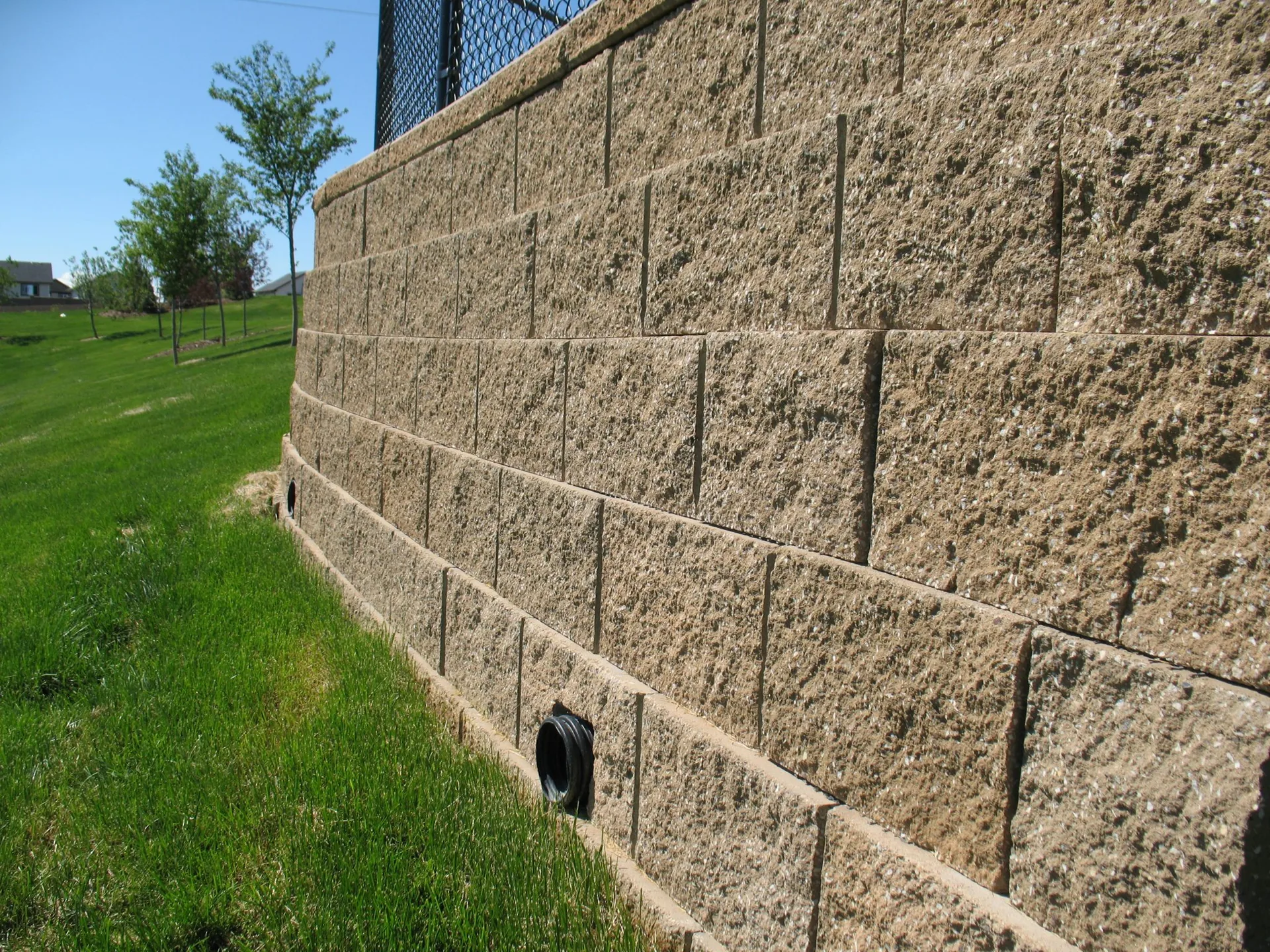 Tall split-face block retaining wall with visible drainage outlets along a grassy slope