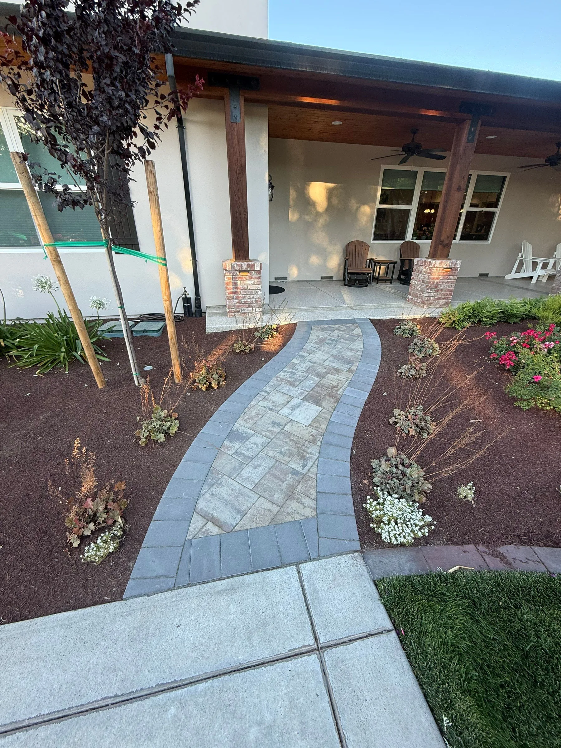 Curved paver walkway leading through planted garden beds to a covered patio