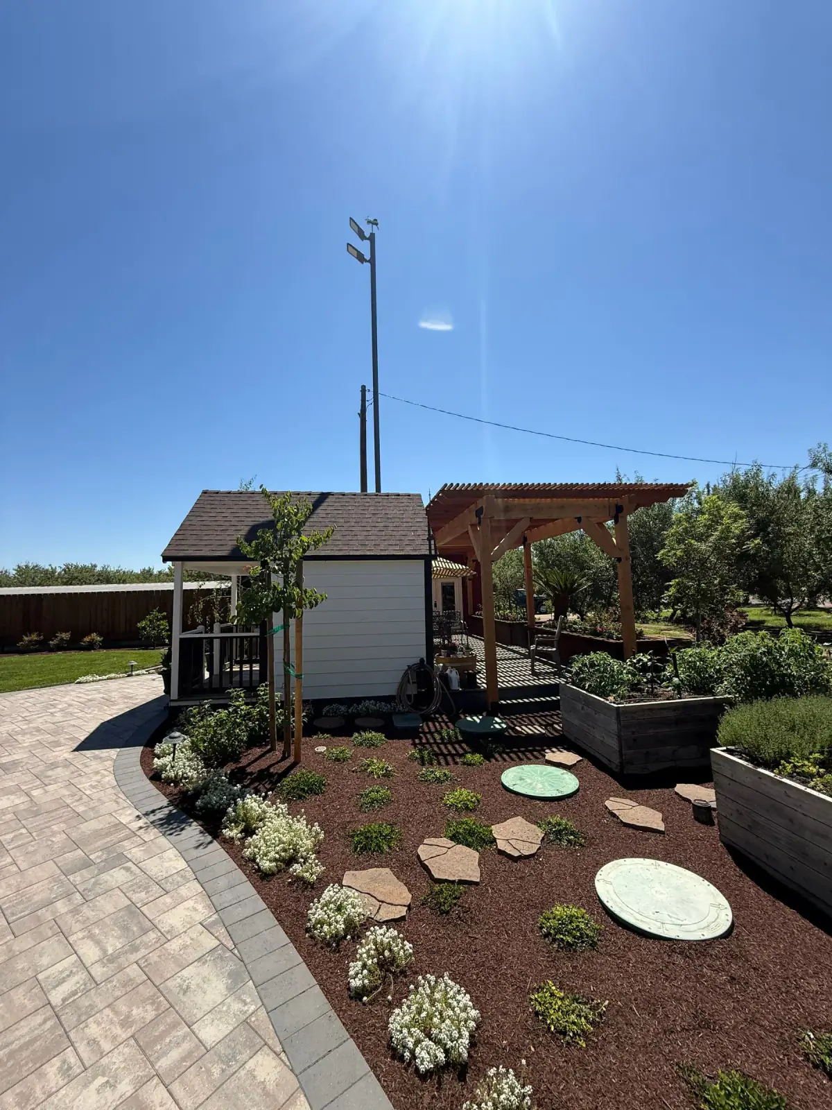 Backyard paver paths, raised planters, and mulched beds around a shed and pergola
