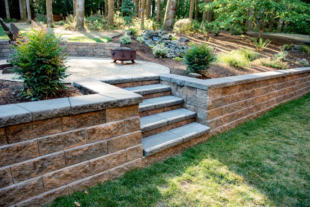 Block retaining wall with steps leading to a landscaped fire pit area.